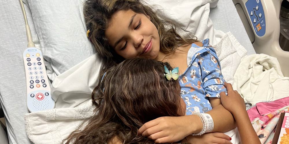 Reggie, a young kidney patient, hugging her sister from her hospital bed.