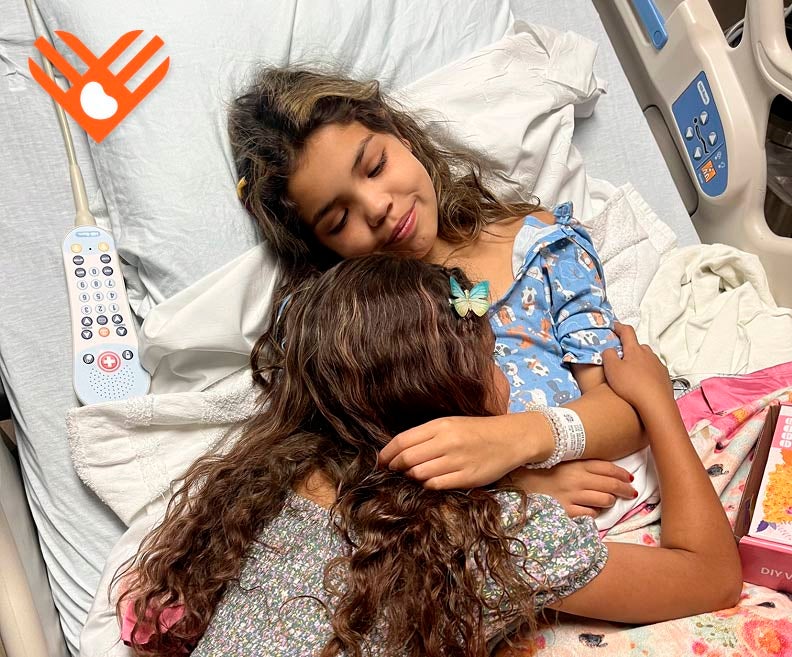 Reggie, a young kidney patient, hugging her sister from her hospital bed. Giving Tuesday heart logo.