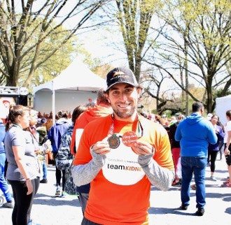 A man wearing an orange team kidney tshirt smiles while holding a race medal