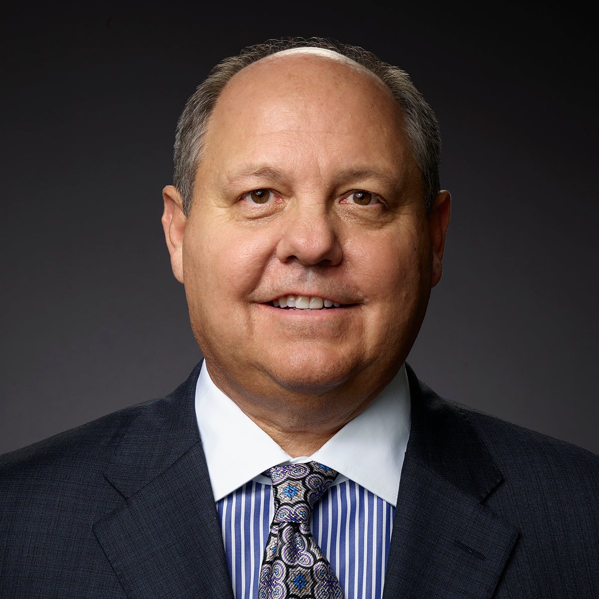 Rick Taylor, a white man in a dark blue suit wearing a paisley tie over a blue and white striped shirt. He is smiling at the camera