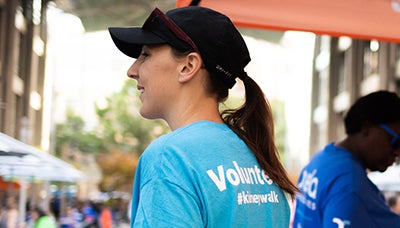 a woman wearing a baseball cap and a blue shirt with the word Volunteer printed on the back. She is volunteering at a Kidney Walk