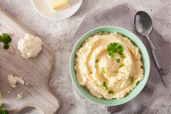 Bowl of mashed cauliflower with whole cauliflower on cutting board next to it.