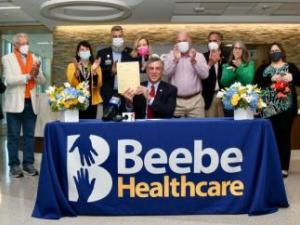 Delaware Governor John Carney sits at a table with a royal blue table cloth that has "Beebe Healthcare" written on the front of it. He is holding up the signed Living Donor Protection Act bill. Eleven people stand behind him in a semicircle.