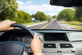 Unas manos sujetan el volante de un coche, que circula por una carretera soleada bordeada de árboles y otros vehículos bajo un cielo azul.
