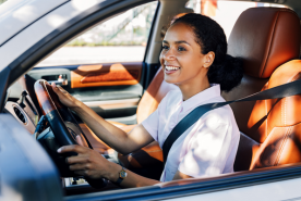 Una mujer sonriente, vestida con una camisa blanca, conduce un coche con asientos de cuero marrón; lleva puesto el cinturón de seguridad y sujeta el volante con las manos.