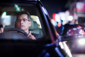 Un hombre con traje y gafas conduce un coche por la noche; a través del parabrisas se ven las luces difuminadas de la ciudad y sus reflejos.