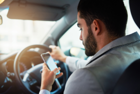 Un hombre dentro de un coche sostiene un smartphone con una mano y el volante con la otra; lleva una camisa gris claro.