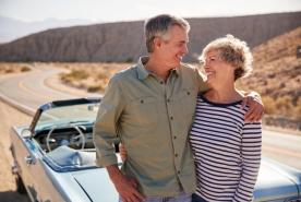 Una pareja de personas mayores sonríe junto a un descapotable azul de época en una sinuosa carretera del desierto, con unas colinas al fondo.