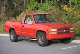 Una camioneta Chevrolet de color rojo brillante, con neumáticos grandes, está aparcada en una calle, rodeada de árboles y arbustos.