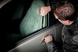 Un hombre con una chaqueta de cuero negra baja la ventanilla del coche mientras sujeta la manilla de la puerta; en el interior del vehículo se ve una manta de color verde claro.