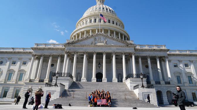 image is a picture of the United States Capitol Building. A group of NKF advocates wearing orange scarves stand on the building's steps holding an American flag.
