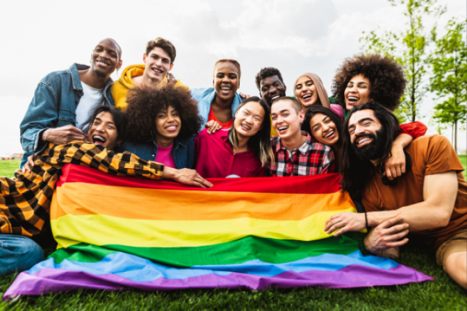 Group of happy adults hugging while sitting on the ground, holding a large LGBTQIA flag