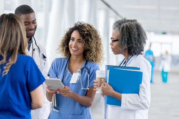 Four healthcare professionals—two nurses in blue scrubs, two doctors in white coats—gather in a hospital hallway, discussing with tablets and folders.